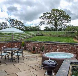 An outdoor patio with a round table and four chairs under a white umbrella a hot tub a small grill potted plants and a grassy area with wooden fencing at Owl Cottage in Napton-on-the-Hill near Southam