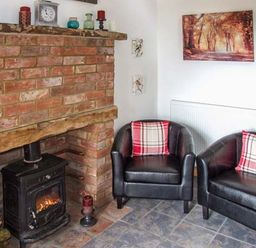A room with a brick fireplace a black wood stove two black leather armchairs with red plaid cushions and wall decorations at Owl Cottage in Napton-on-the-Hill near Southam