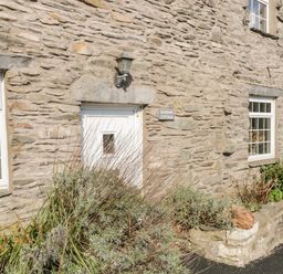 Stone exterior wall of Duddon Cottage with white door and windows surrounded by shrubs and plants at Broughton-in-Furness