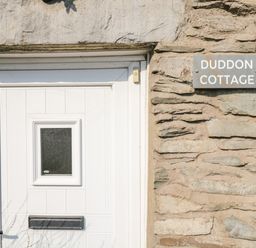 A white door with a small square window and a mailbox on a stone wall with a sign that reads Duddon Cottage at Broughton-in-Furness