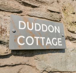 A close up of a gray metal sign with white letters saying Duddon Cottage mounted on a stone wall at Duddon Cottage in Broughton-in-Furness