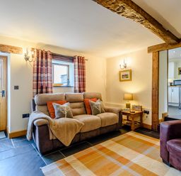 A living room with a sofa and armchair near a wooden door and window with plaid curtains at The Stables in Llanfair Caereinion near Welshpool