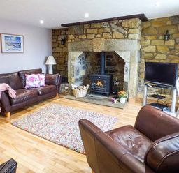 A living room with brown leather sofas a stone fireplace with wood stove and a television on a stand at Sandy Knowes in Low Hauxley near Amble