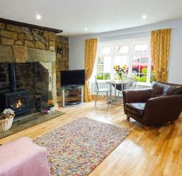 A living room with a stone fireplace wood burner a television leather armchairs a dining table with flowers and yellow curtains at Sandy Knowes in Low Hauxley near Amble