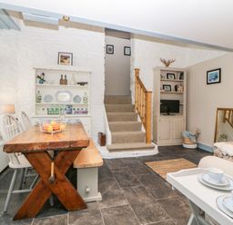 A dining and living area with a wooden table, chairs, a bookshelf, stairs, and a small TV at The Old Dairy in Maker near Millbrook