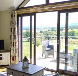A living room with a wooden coffee table a small lantern a television on a white cabinet and glass doors leading to a balcony with rattan chairs at Black Mountain View in Llanafan Fawr near Builth Wells