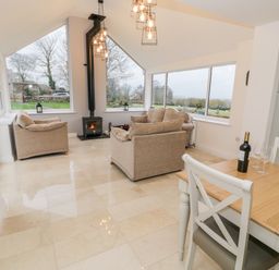 A sitting area with beige sofas and a wood stove surrounded by large windows next to a dining table with chairs and a bottle of wine at Ty'n Lon Bach in Llandwrog