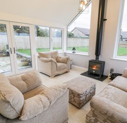 A sitting room with beige armchairs a floral patterned sofa an ottoman a wood burning stove and large windows at Ty'n Lon Bach in Llandwrog