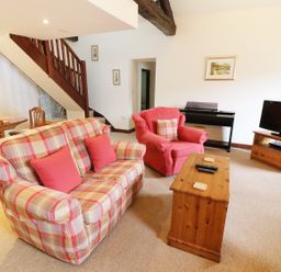 A living room with a plaid sofa and red armchair a wooden coffee table a TV on a wooden stand a piano and dining table near stairs at Kingfisher Barn in Staveley near Windermere