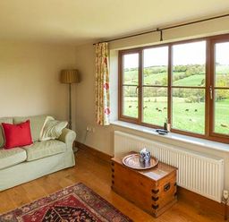 A living room with a sofa a floor lamp a wooden chest a window with curtains and a view of green fields at Glebe Farm Cottage in Llandegveth near Cwmbran