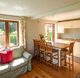 A living area with a green sofa and red cushion next to a wooden dining table with four chairs and a kitchen in the background at Glebe Farm Cottage in Llandegveth near Cwmbran