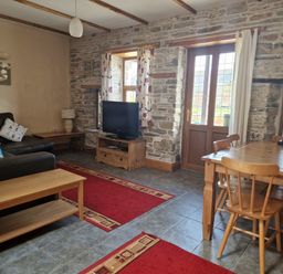 A living room with a black sofa, wooden coffee table, TV on a wooden stand, dining table with chairs, stone walls, and red rugs at Kingfisher Cottage in Llanboidy