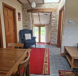 A hallway with a wooden dining table and chairs a blue armchair with a cushion red rugs on a tiled floor wooden doors a stone archway and glass doors leading outside at Kingfisher Cottage in Llanboidy