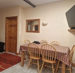 A dining area with a table covered with a checkered cloth and six wooden chairs with a wall-mounted TV and a wooden door at Puffin Cottage in Llanboidy