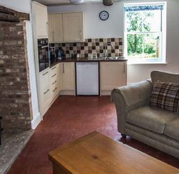 A small kitchen area with stove and cabinets next to a stone fireplace and living room with sofa and cushions at Cotswold View Clifford Chambers near Stratford-upon-Avon