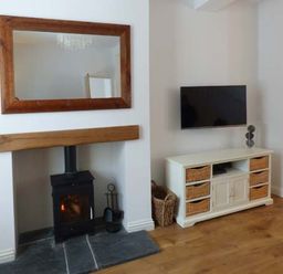 A room with a wood burning stove under a wooden mantel and a wall mounted TV above a cabinet with wicker baskets at Silversands Cottage in Amble-by-the-Sea