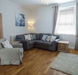A living room with a gray corner sofa a patterned armchair wooden floor and a window with curtains at Silversands Cottage in Amble-by-the-Sea