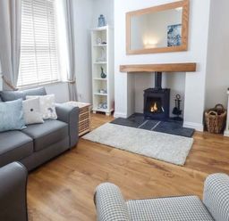 A living room with a gray sofa cushions a wood burning stove and shelving at Silversands Cottage in Amble-by-the-Sea