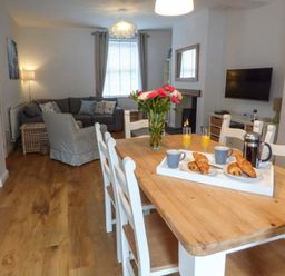 A dining area with a wooden table set with breakfast items and a vase of flowers and a living room with a sofa and armchair at Silversands Cottage in Amble-by-the-Sea