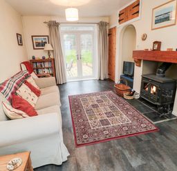 A living room with a beige sofa red and patterned cushions a wooden bookshelf a patterned rug a fireplace and glass doors leading outside at Buntys Place in Chatton near Wooler