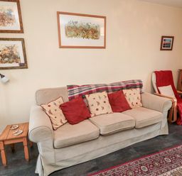 A living room with a beige sofa with red and patterned cushions a wooden side table a standing lamp and framed artwork on walls at Bunty's Place in Chatton near Wooler