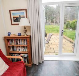 A room with a wooden bookshelf a red chair a lamp and a glass door leading to a patio and garden at Bunty's Place Chatton near Wooler