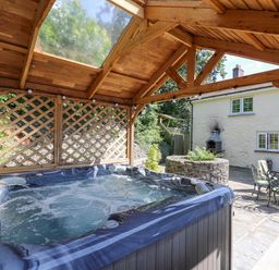 An outdoor hot tub under a wooden pergola next to a stone patio with metal table and chairs and a white house in the background at Penuwch Fach in Aberystwyth