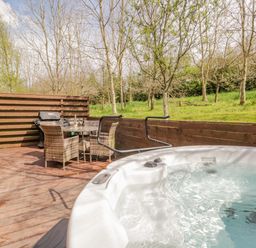 An outdoor wooden deck with a hot tub a dining table with four wicker chairs and a barbecue grill surrounded by trees at Cartmel Lodge in Allithwaite near Grange-over-Sands