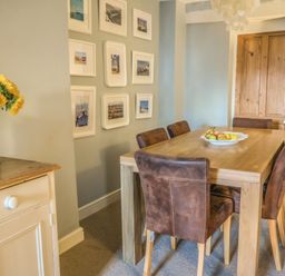 A dining room with a wooden table and six brown chairs a wooden door and framed pictures on the wall at Craft Cottage in Beer near Seaton