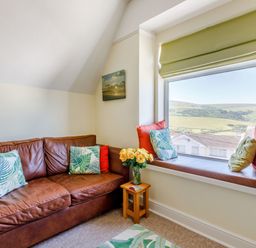 A sitting area with a brown leather sofa, cushions, a small wooden side table with flowers, and a window seat with cushions overlooking a landscape at Glenwood in Woolacombe