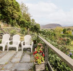 Three white plastic chairs on a stone patio with flowers in wooden planters and a garden with shrubs and mountains in the background at Ty Gwynn in Tanygrisiau