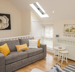 A sitting area with a grey sofa and yellow cushions near white tables and a window with blinds at The Store Rooms in Whitby