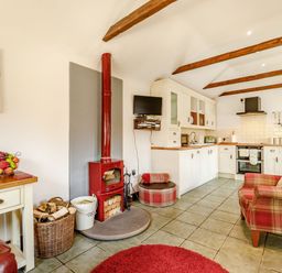 An open plan kitchen and living area with white cabinets a red wood stove and a plaid armchair at Willow Cottage in Ulrome near Skipsea