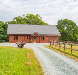 A wooden house with a gravel driveway and fenced grassy area surrounded by trees at Hampton Lodge in Ellesmere