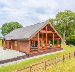 A wooden cabin with a gray roof and a covered porch in a grassy area with trees at Hampton Lodge in Ellesmere