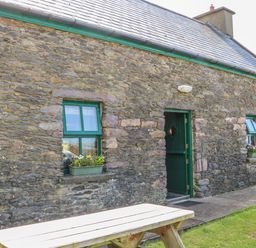 A stone building with green framed windows and a green door with flower boxes under the windows and a wooden picnic table outside at An Nead in Ballinknockane near Dingle County Kerry