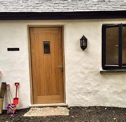 A cottage entrance with a wooden door a wall light an open black framed window and children's toys on the ground at Vine Cottage in St Ewe near Sticker