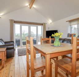 A dining area with wooden table and chairs a vase with flowers a living room with leather sofas a television and french doors at Vine Cottage in St Ewe near Sticker