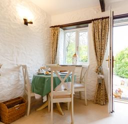 A dining area with a round table covered with a green tablecloth and four chairs near a window and an open door at Poldark Cottage in East Taphouse