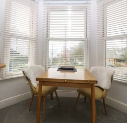 A small dining area with a wooden table and two chairs next to three windows with blinds at Y Castell Apartment 2 in Criccieth