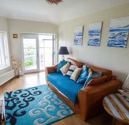 A living room with a brown sofa with blue cushions and a patterned rug and a small wooden table with a model sailboat at Ty'r Enfys Bach - Upper in Trearddur Bay