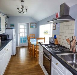 A kitchen with white cabinets black countertops a gas stove wooden dining table and chairs at Ty'r Enfys Bach - Upper in Trearddur Bay