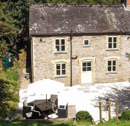 A stone house with four windows and a door beside a patio with outdoor furniture and an umbrella at Ploony Cottage in Bleddfa
