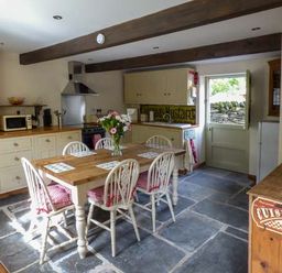 A kitchen with a wooden table and six chairs with cushions a vase of flowers on the table cupboards a microwave a stove and a door leading outside at Ploony Cottage in Bleddfa