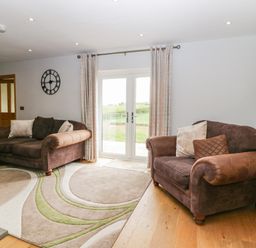 A living room with two brown sofas and patterned rug near glass doors with curtains at Primrose Cottage in Aldingham