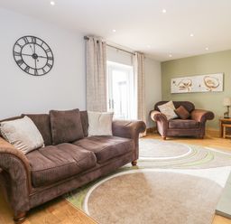 A living room with two brown sofas a wall clock on a white wall a wooden door and a large wall painting at Primrose Cottage in Aldingham