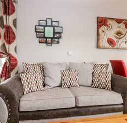 A living room with a sofa with patterned cushions a floral painting on the wall and red and beige curtains at Daisy Cottage in Aldingham