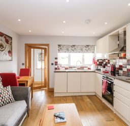 A kitchen with white cabinets red and white tiled backsplash a wooden floor a gray sofa with patterned cushions and a wooden table with red chairs at Daisy Cottage in Aldingham