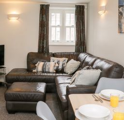 A living room with a brown leather sectional sofa with pillows, a matching ottoman, a television on a stand, and a dining table set with plates and glasses of orange juice at Razorbill Cottage in Whitby