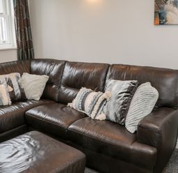 A corner of a living room with a dark brown leather sectional sofa and several textured pillows at Razorbill Cottage Whitby